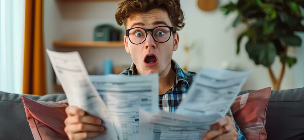 Shocked young man holding papers with a bill and role showing high energy bills at home, surprised black guy sitting in the living room reading scattered paper documents feeling poor