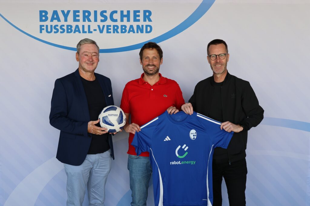Bild mit drei Männern, Mann in der Mitte hält Trikot in den Händen.