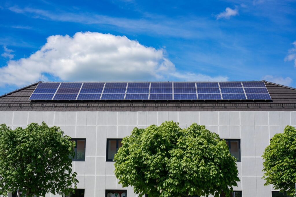 Ein modernes Gebäude mit Photovoltaik-Solarzellen auf dem Dach, drei grüne Bäume davor, unter einem blauen Himmel mit vereinzelten Wolken, das seinen Bewohnern nachhaltigen Mieterstrom bietet.